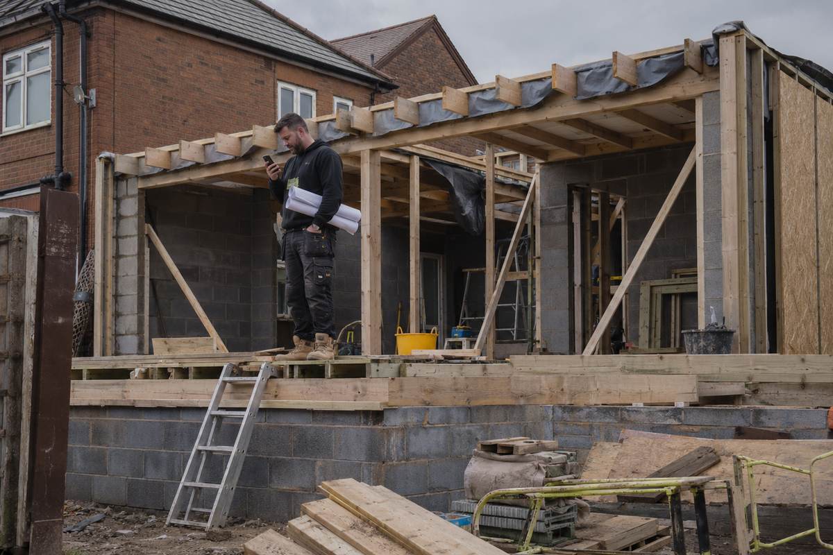 A builder standing on a half-finished extension with rolled plans under one arm, checking a phone.
